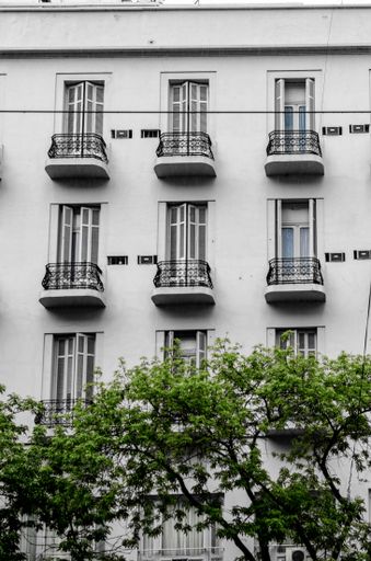 A vertical shot of a white building with balconies in rows with green trees below