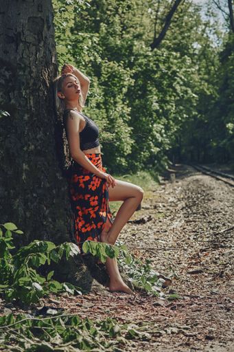 A vertical shot of an attractive blonde female with a floral dress leaning against a tree.