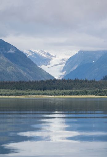 A vertical shot of clear lake with green forests and snowcap mountains in the background