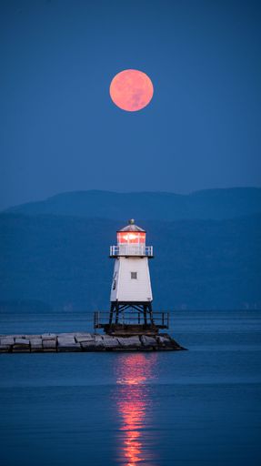 A mesmerizing view of a Harvest Moon Setting over Lake Champlain and a lighthouse, in Burlington with a silhouette of a mountain in the background