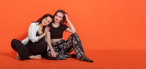 Two young brunette girls in casual clothes, sit on floor on orange background, smile and look at camera. Mother rested head on daughter's shoulder.