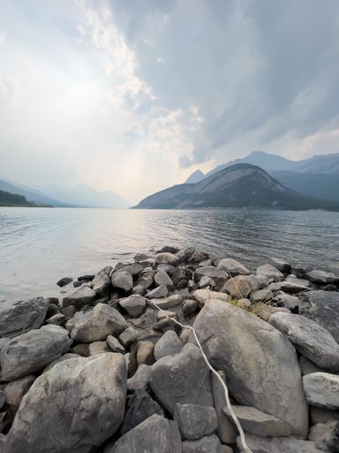 A lovely view of huge rocks by the shore of a serene lake surrounded by hills under a cloudy sky