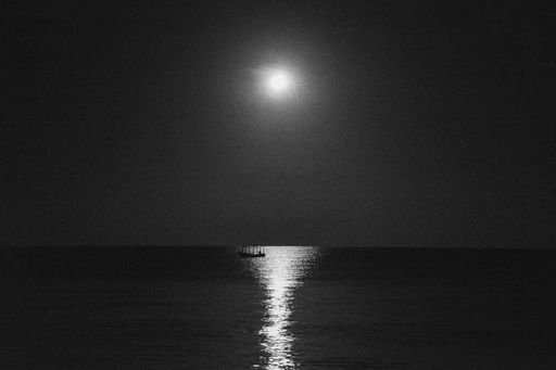 A grayscale shot of a boat in the calm sea with the reflection of the moon at night