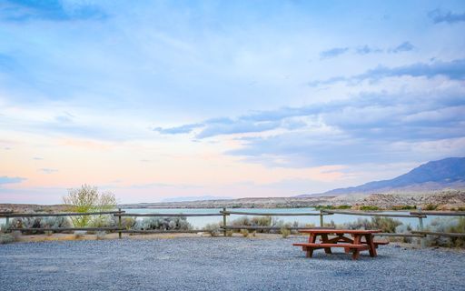 A picnic table is set off a gravel road overlooking a lake and large colorful dramatic cloudy skies.