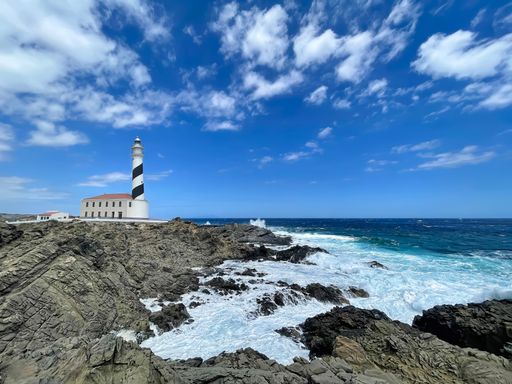Favaritx lighthouse with its tower painted with a blue and white spiral on a rocky cliff with rough sea, Menorca, Balearic Islands, Spain