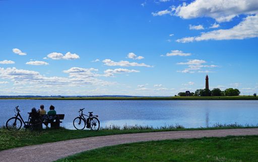 An image of a road for bicycles, two people sitting on a bench in front of a water surface