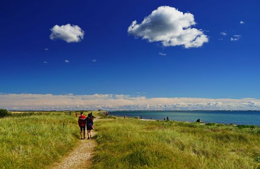 An image of a road surrounded with grass and people walking on it, the Baltic Sea on the right and a beautiful sky in Germany