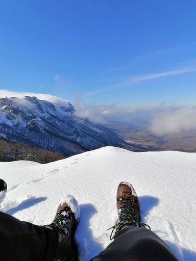 A vertical shot of man's feet sitting on the edge of a hill with  snow-covered mounts in front of him