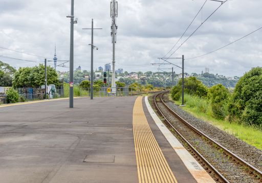A wide shot of a train track near an asphalt road surrounded by green trees in Auckland, New Zealand