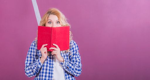 Young girl blonde covered half her face from below with red book and looks in surprise with big eyes at camera. Pink rear background.