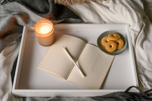 A top view of burning aroma candle, open notebook, cookies and pencil on white wooden tray
