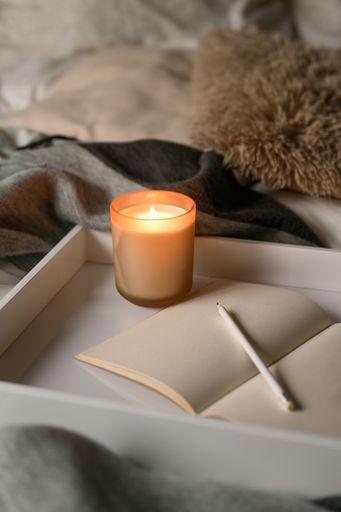 A vertical shot of burning aroma candle, open notebook and pencil on white wooden tray