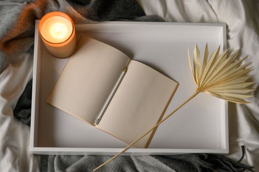 A top view of burning aroma candle, open notebook and pencil on white wooden tray