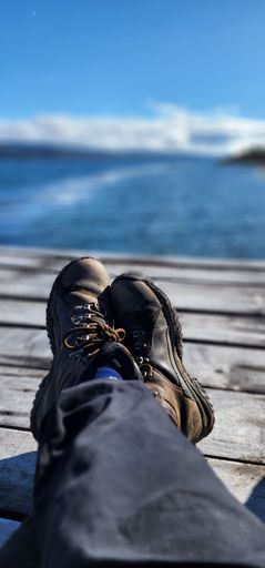 A vertical shot of the feet of a human with dark brown shoes sitting on the dock with the background of a sea