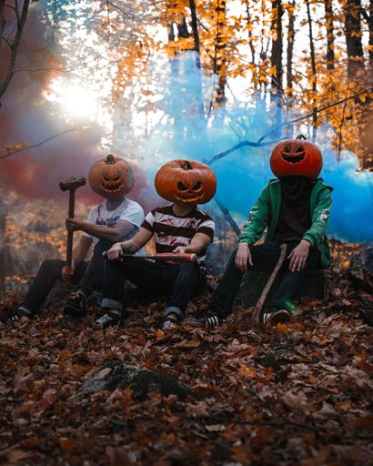 A three guys wearing pumpkins on their heads and sitting in a park during fall with smoke in the background