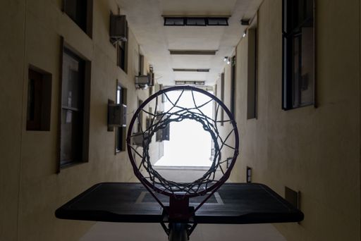 A low angle of a basketball hoop illuminated in the dark, with tall buildings in the background