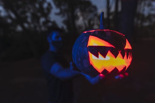 A closeup shot of a Halloween pumpkin with a scary face in the hand of a young male wearing a mask