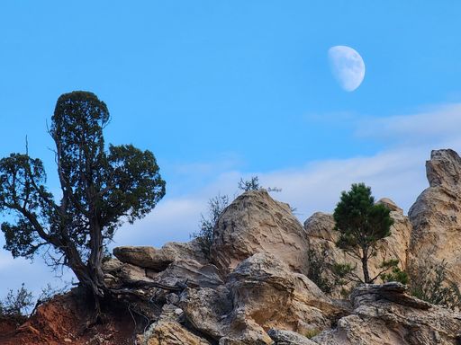 A low angle shot of a half full moon in a blue sky over rocky mountains