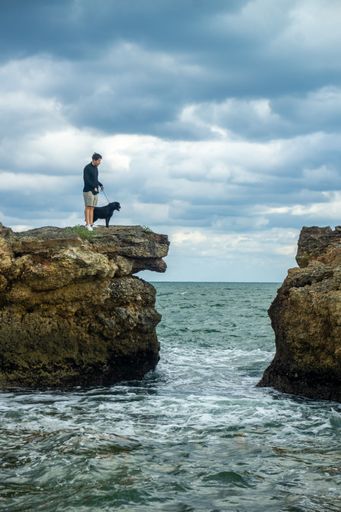 A vertical shot of a male with his dog standing on the rock in the sea