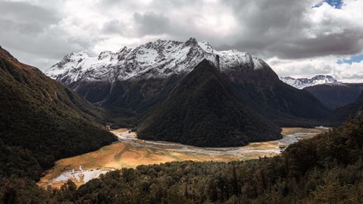 A panoramic shot of partially snow-covered mountains with a pond and stream under the cloudy sky, New Zealand