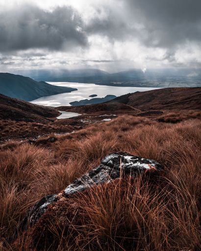 A vertical shot of a grassy mountain range with a river under a cloudy sky in New Zealand