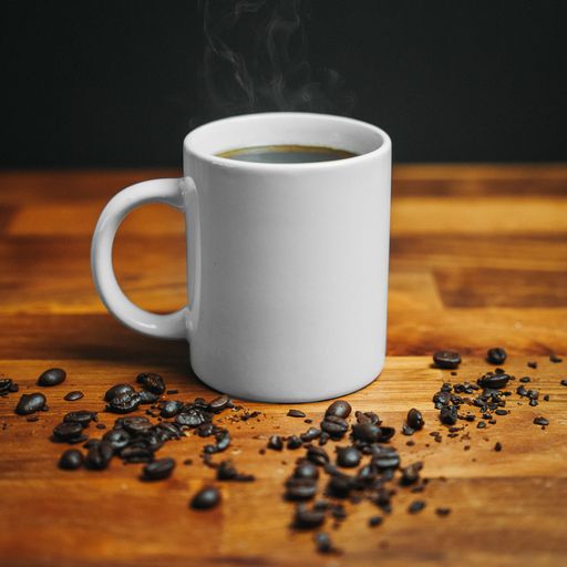 A white mug of steaming hot coffee surrounded by coffee beans on a wooden table