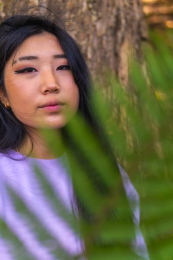 A vertical shot of a beautiful Chinese girl's portrait, posing near trees behind a leaf, outdoors during daylight