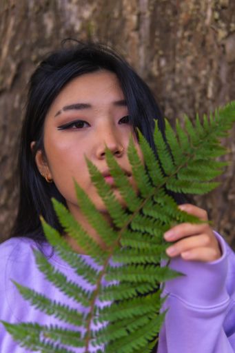 A vertical shot of a beautiful Chinese girl's portrait, posing near trees holding a leaf, outdoors during daylight