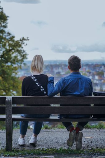 A vertical of a Caucasian couple sitting on a bench, seen from behind in a park overlooking the city
