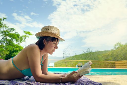 A close-up shot of a female reading a book in the sun near a swimming pool