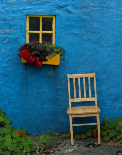 A vertical shot of yellow window flower box on blue old wall and wooden chair under them in Ireland