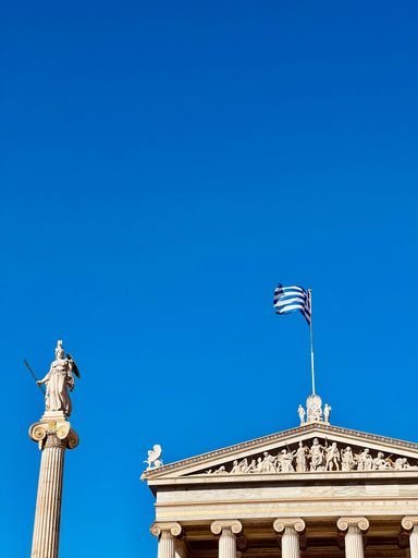 The greek flag on the top of Academy of Athens university on blue sky background in Athens, Greece
