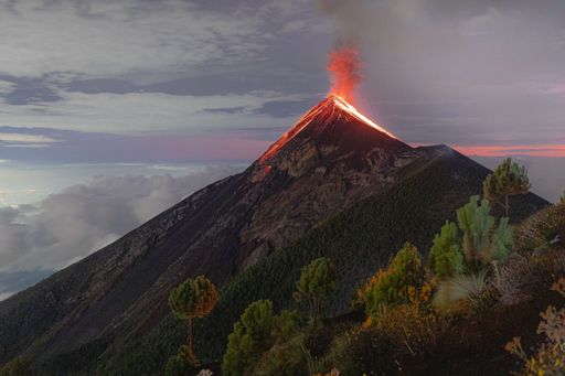 An aerial view of Volcano de Fuego, Guatemala, erupting, covered with grass and trees, with smoke and lava around the top