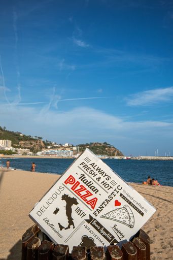 Pizza box at home in the foreground with the background of the beach of Blanes with people relaxing in it under a beautiful blue summer sky.