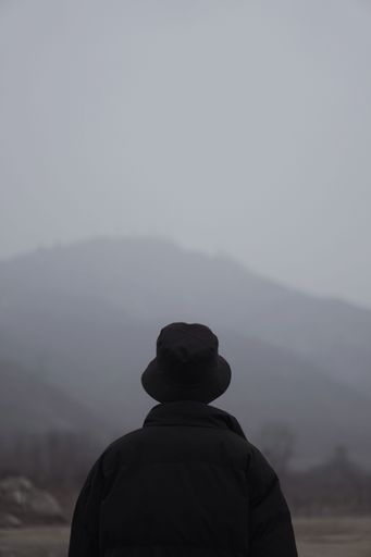 A vertical shot of a male in black clothes and hat seen from behind looking at mountains in fog