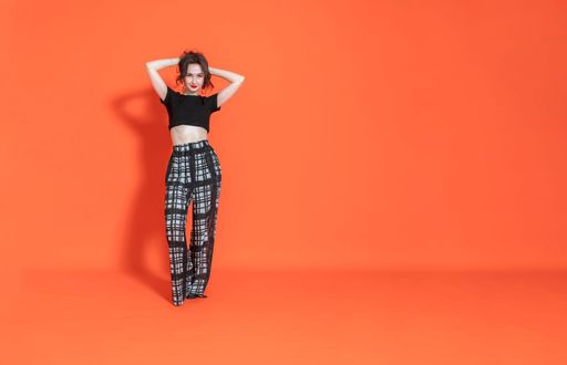 Cheerful young brunette girl in black and white trouser casual suit stands on orange background, hands are raised and behind head, looks into camera.