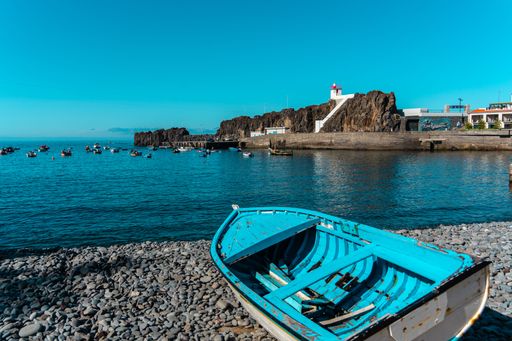 A wooden boat on the rocky beach in Madeira, Portugal, with boats in the rocky pier in the background, on a sunny day