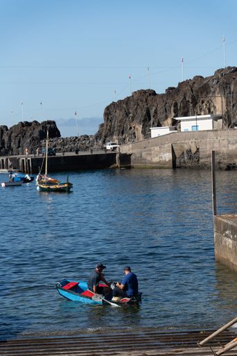 Two men in a small boat, rowing in Madeira, Portugal, with boats in the rocky pier in the background, on a sunny day