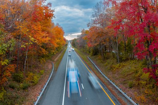 A bird's eye view of cars driving fast on a highway between autumn-colored trees