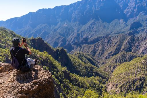 A young male resting after trekking at the top of La Cumbrecita sitting in the natural viewpoint and taking a photo with the mobile