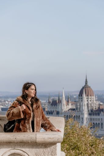 A vertical shot of a girl standing on a stone balcony with a Budapest cityscape in the background