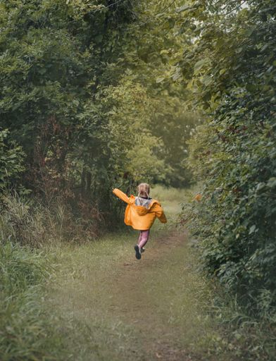 A back view vertical shot of a blond little girl with braids hopping on a dirt road among the forest