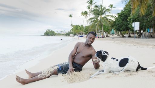 Happy African American male tourist in shorts sitting on sand and petting obedient dog while spending summer weekend day on beach