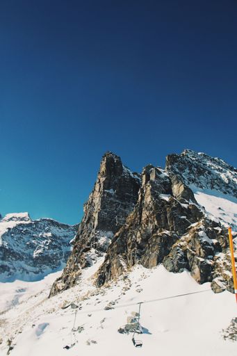 A vertical shot of big rocky mountains covered in snow and a ropeway against the clear blue sky during daytime