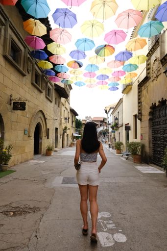 A vertical back view of a female in short walking in an alley with overhead hanging colorful umbrellas