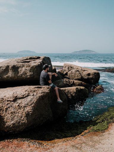 Beautiful landscape shot of a guy sitting on a rock on the beach during a bright sky summer day.