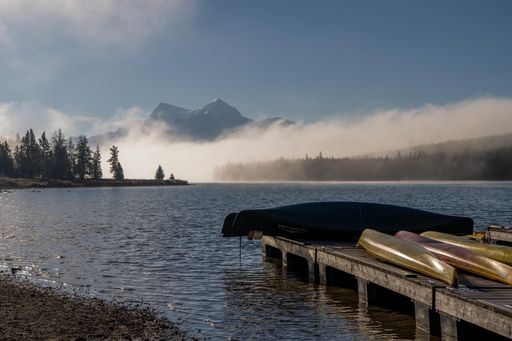 A wooden boardwalk on the seashore with canoes in front of high mountains covered in fog