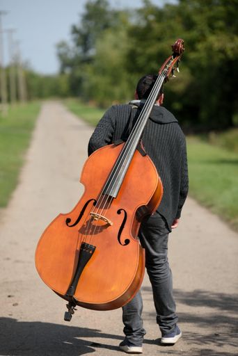 A man with a cello on his back waking in a green park