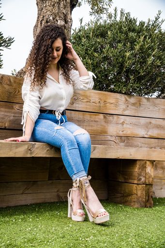 Young woman with curly brown hair, wearing a white shirt, blue jeans and heels, sitting with her feet crossed on a wooden bench with grass and trees