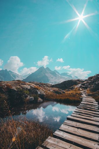 A vertical shot of a narrow wooden passage over a reflective small lake and a mountain range on the horizon